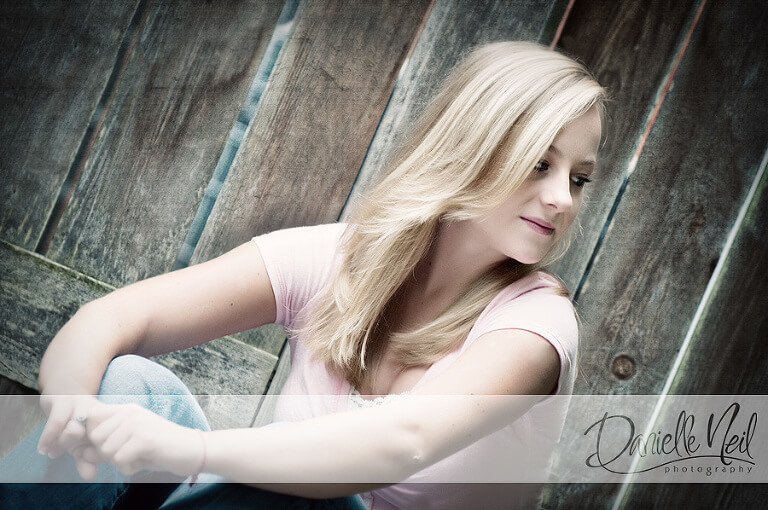 Blonde teen sitting against wooden fence with pink shirt on